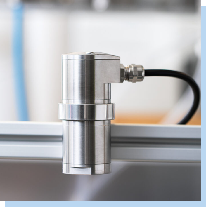 Close-up of the NPS-P2 mounted on a metal rail, with a black cable connected at the top, set against a blurred laboratory or manufacturing background.